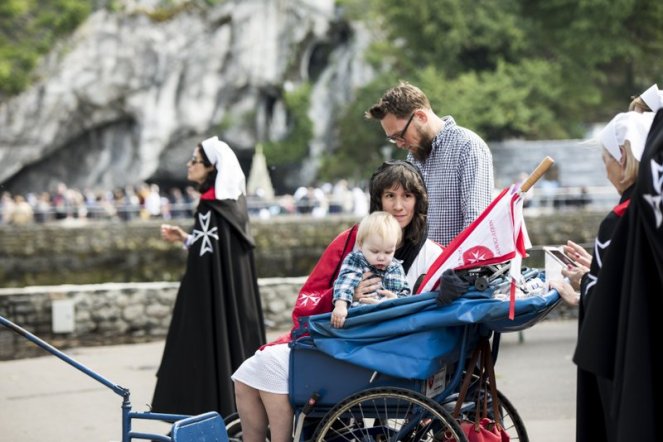 Image: Woman sitting in a blue cart, with a blond infant on her lap. A man walks next to the cart. They are accompanied by two women in white veils and black capes that bear a white eight-pointed cross.