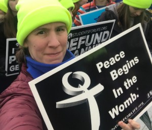 Women taking a selfie, wearing a purple winter coat and neon yellow hat. She holds a sign that reads, "Peace Begins in the Womb."